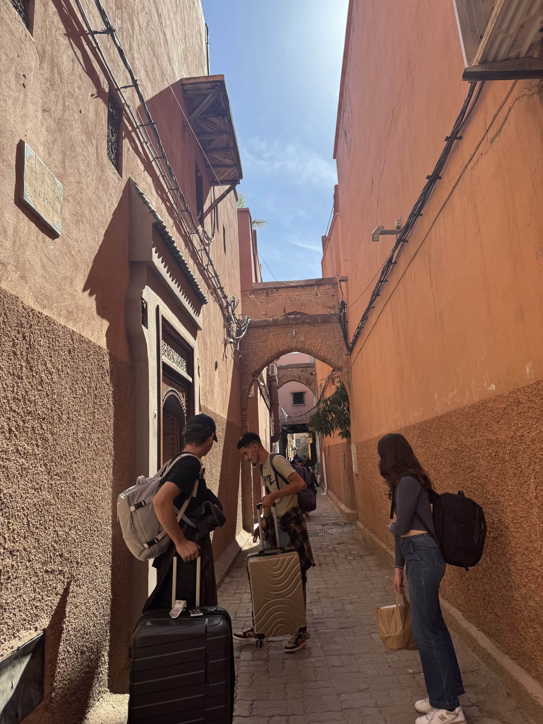 Viajeros con maletas recorriendo un callejón de la medina roja de Marrakech bajo un arco tradicional