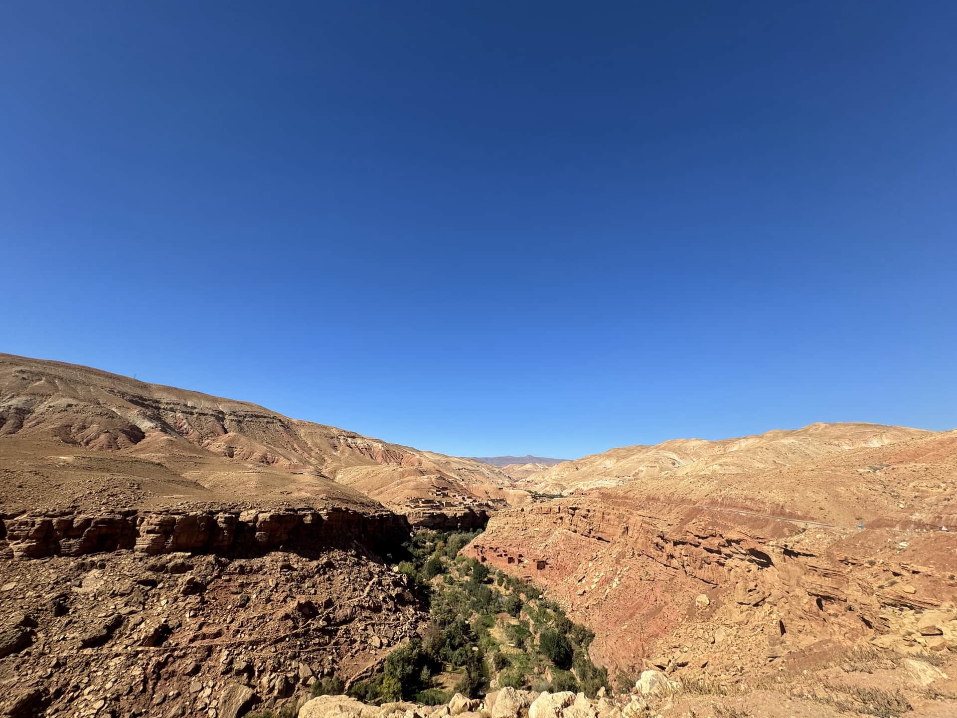 Valle del Dadés desde las alturas con cañón rocoso rojizo, vegetación verde en el fondo y cielo azul despejado, Marruecos