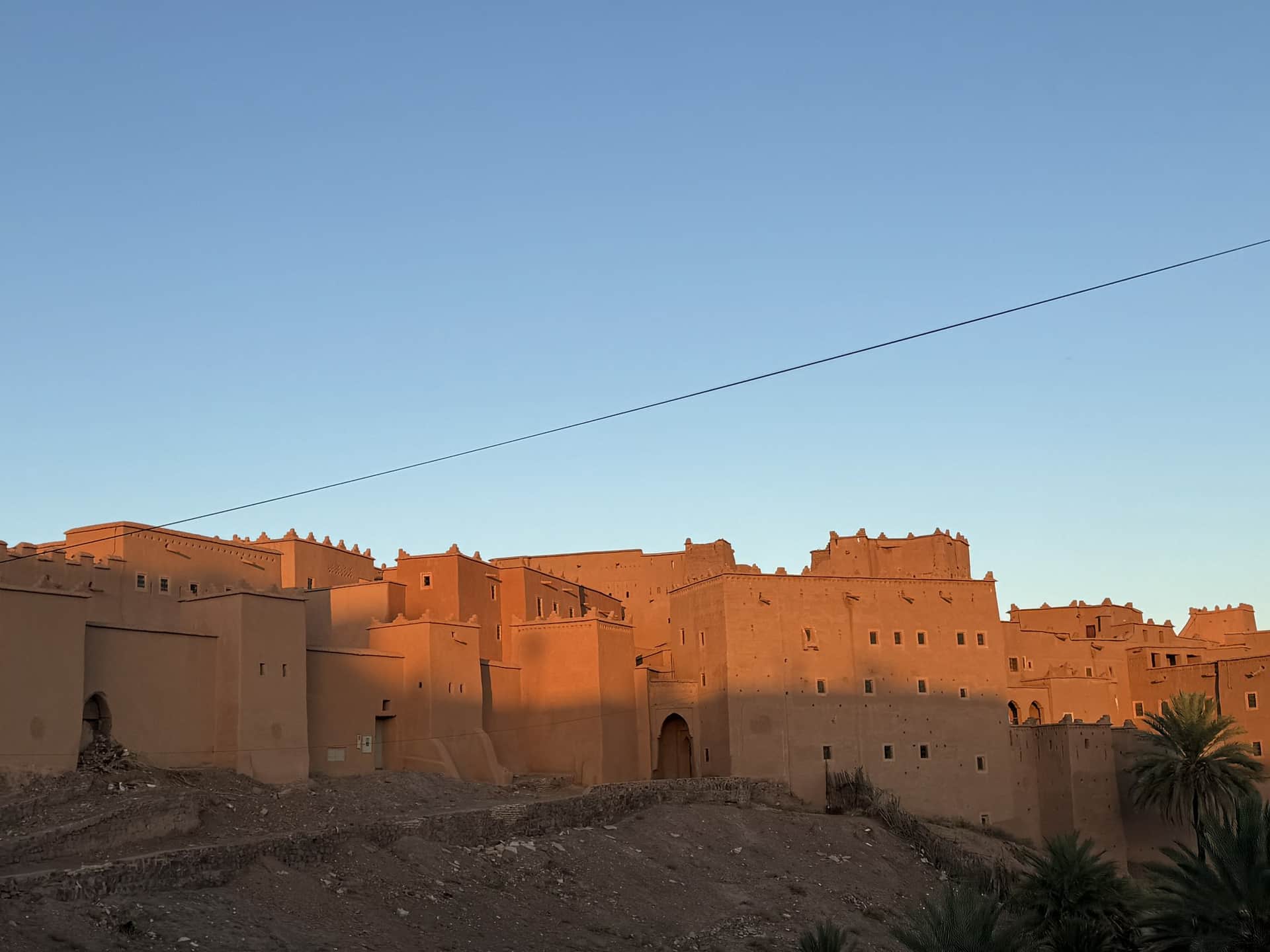 Kasbah Taourirt de Ouarzazate al atardecer, muros de adobe rojizo bajo cielo azul con palmeras