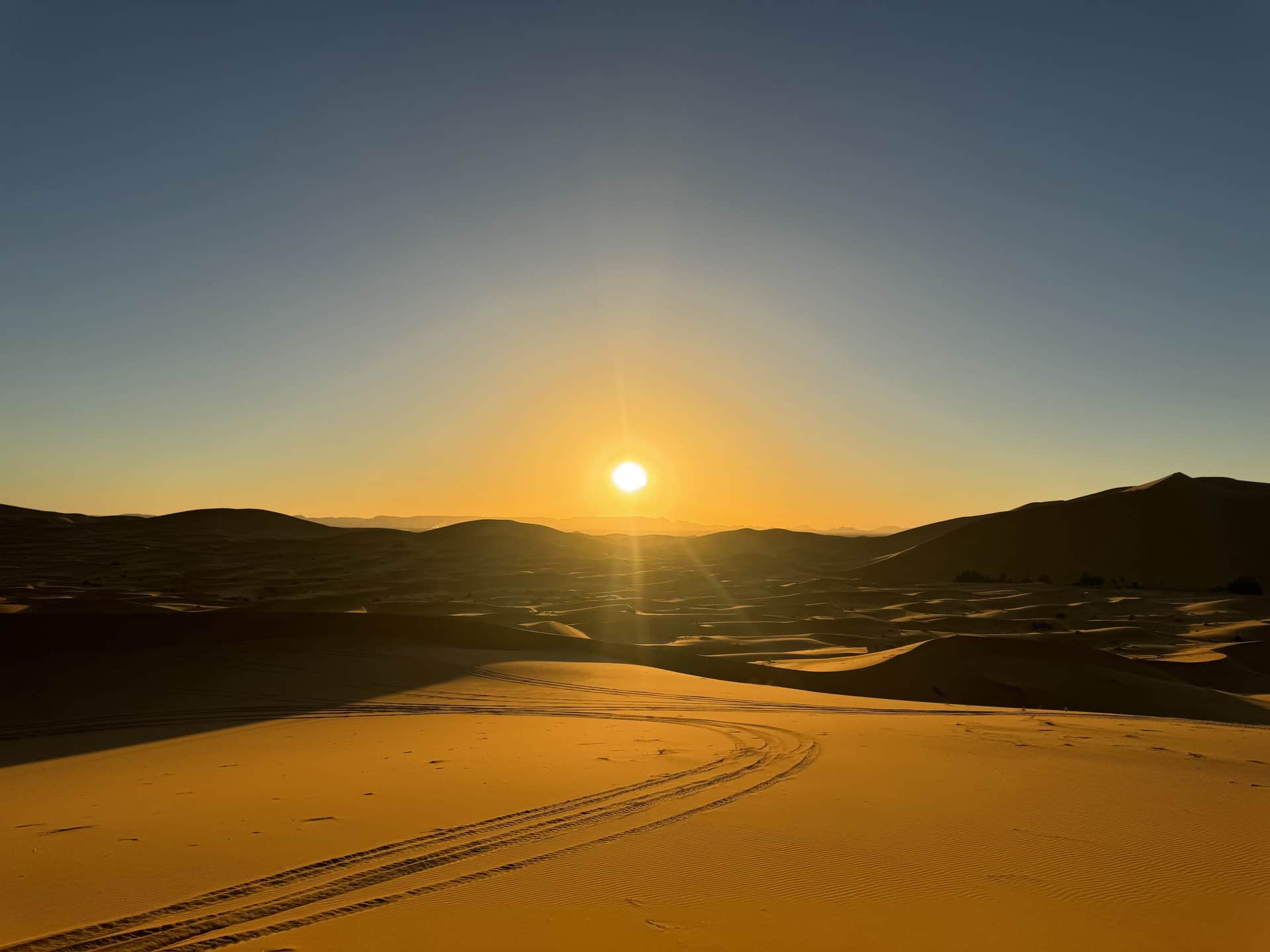 Amanecer dorado sobre las dunas del Erg Chebbi en Merzouga, con huellas de 4x4 en la arena del Sahara