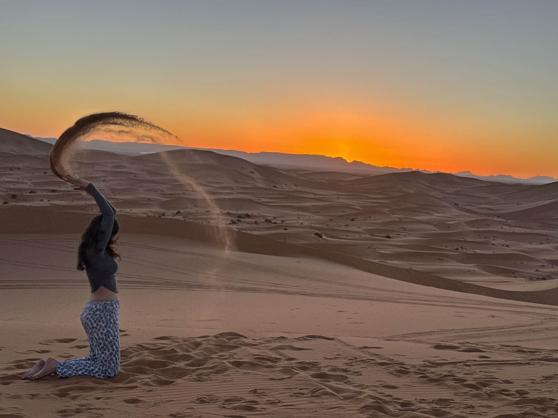 Mujer lanzando arena al atardecer en las dunas del Sahara de Merzouga, cielo naranja intenso
