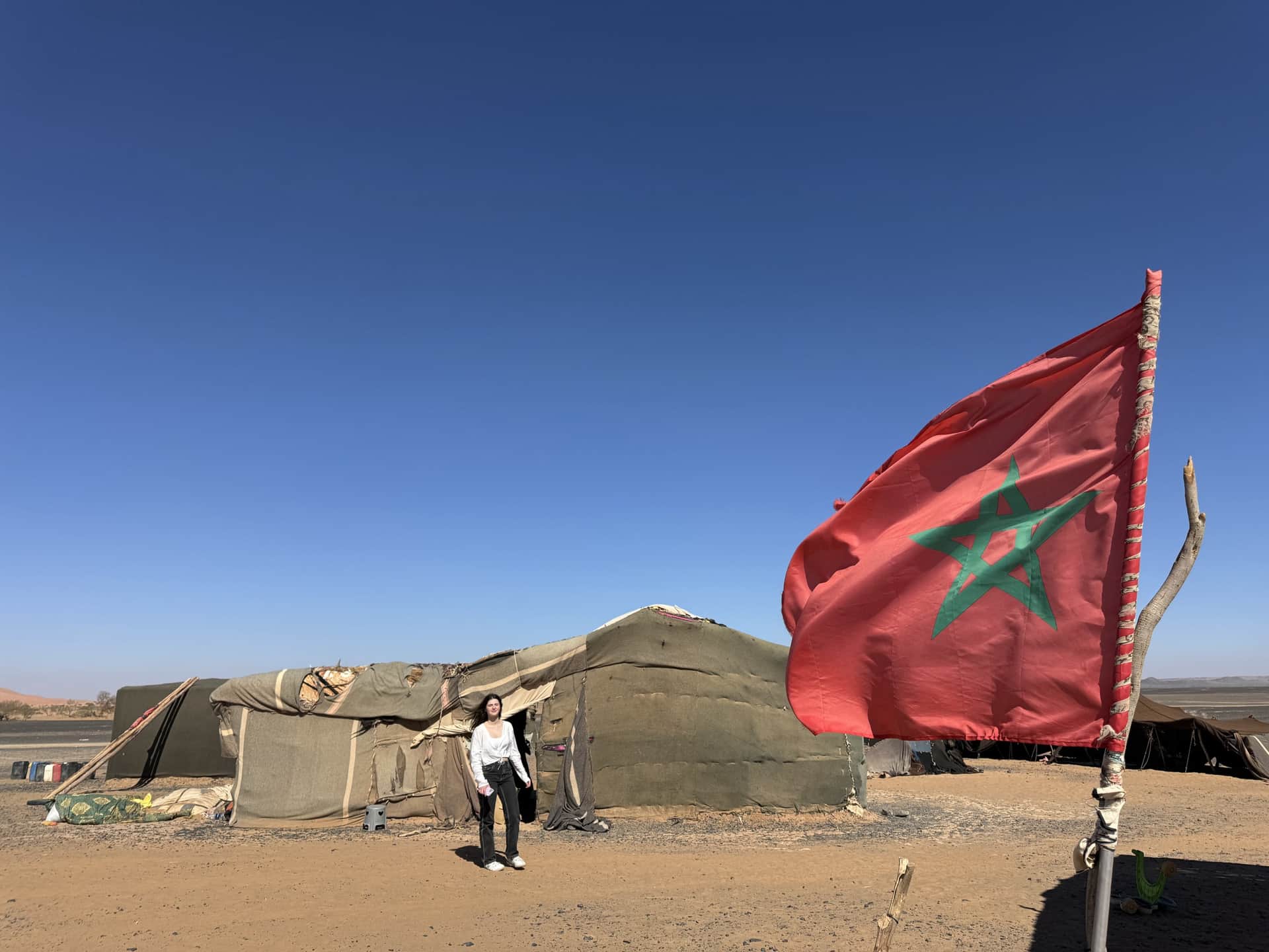Viajera junto a jaima nómada en el desierto de Merzouga con la bandera de Marruecos ondeando bajo cielo azul