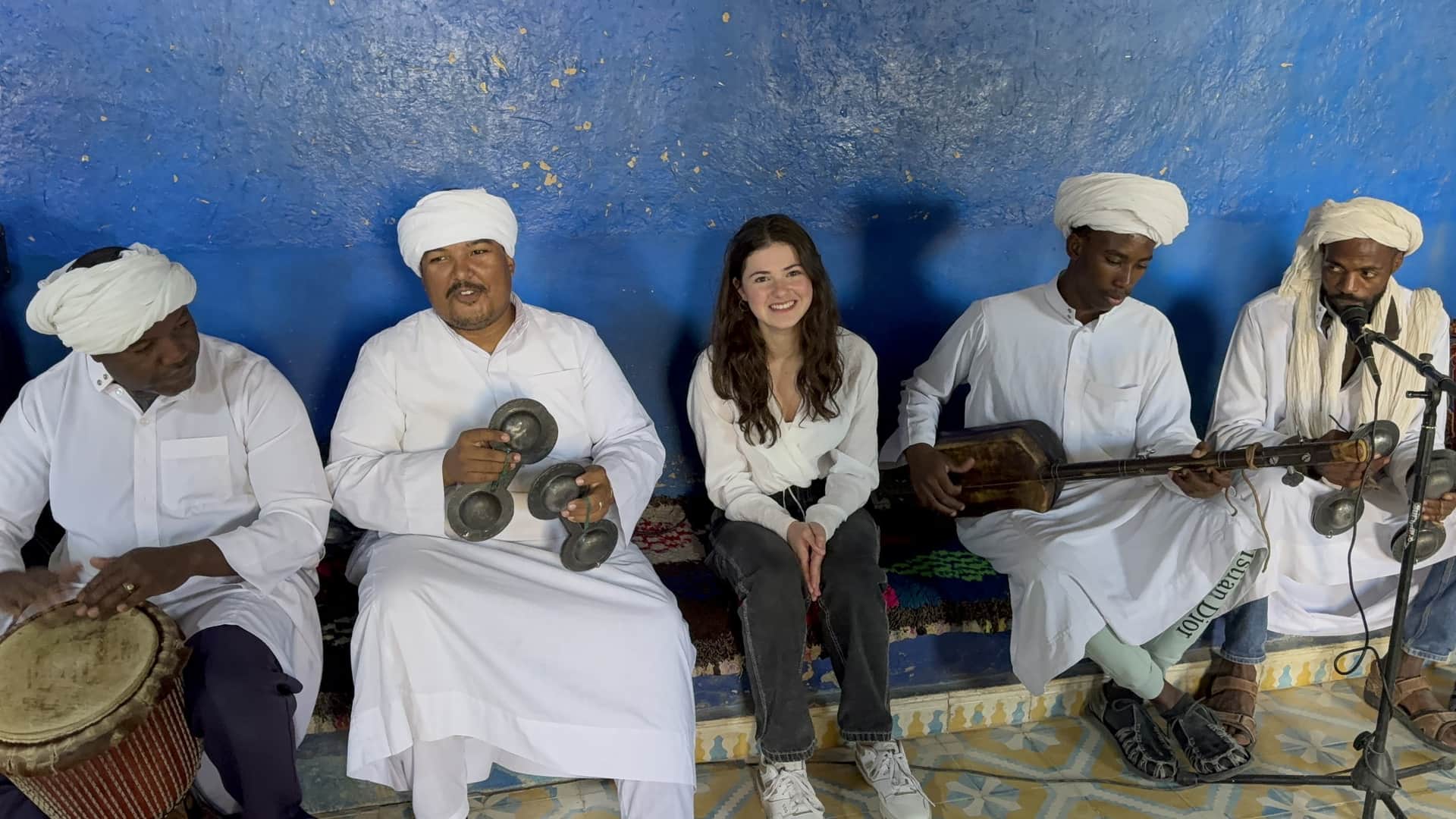 Viajera sonriendo junto a músicos Gnawa en Khamlia, Merzouga, con instrumentos tradicionales y pared azul