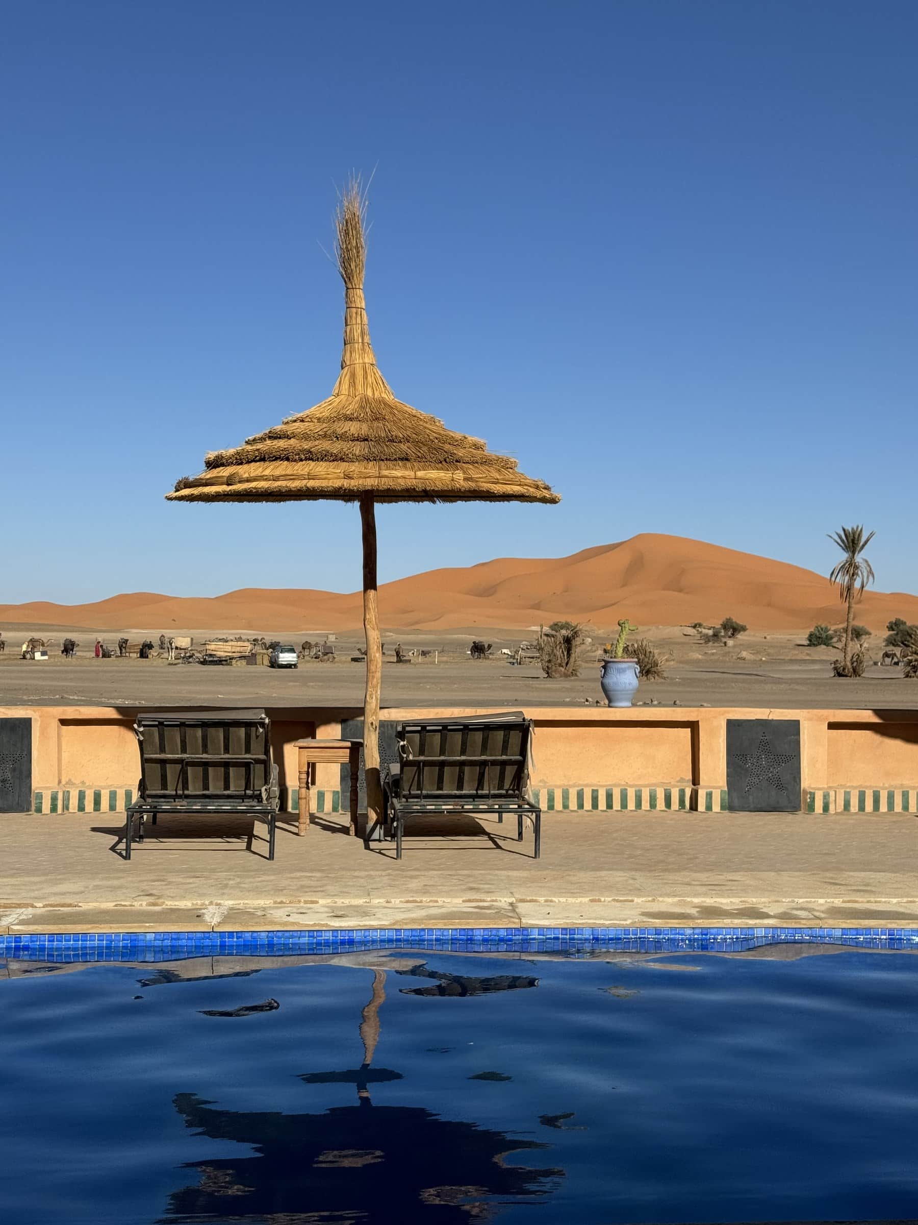 Piscina de hotel en Merzouga con vistas a las dunas del Erg Chebbi y dromedarios al fondo, Sahara marroquí