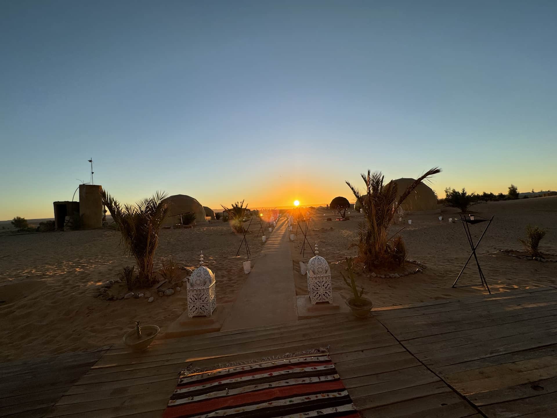 Campamento de lujo en el desierto de Merzouga al atardecer con jaimas, faroles marroquíes y palmeras bajo el sol poniente