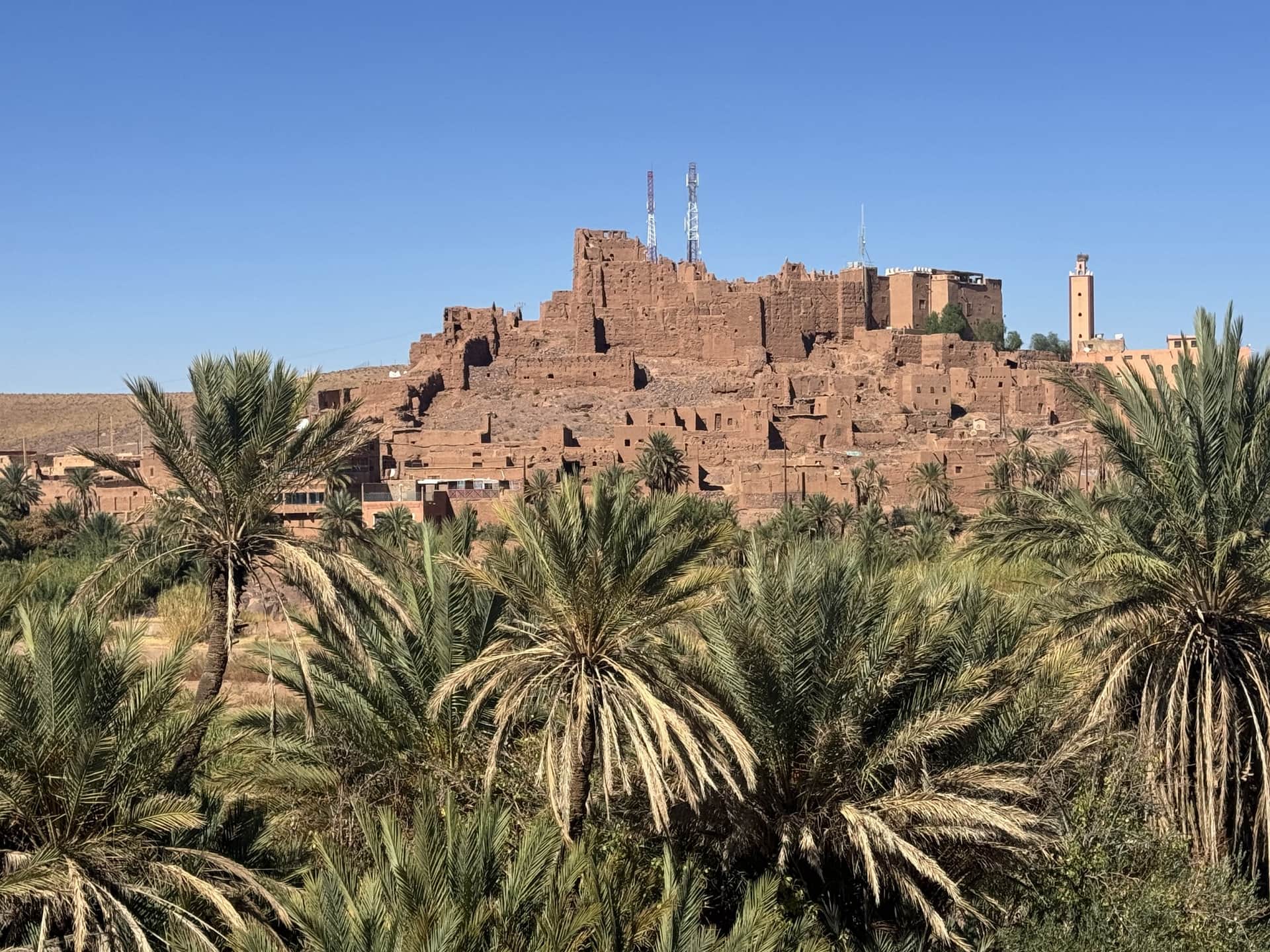 Kasbah de tierra rojiza en ruinas sobre colina rodeada de palmeras datileras bajo cielo azul en el sur de Marruecos