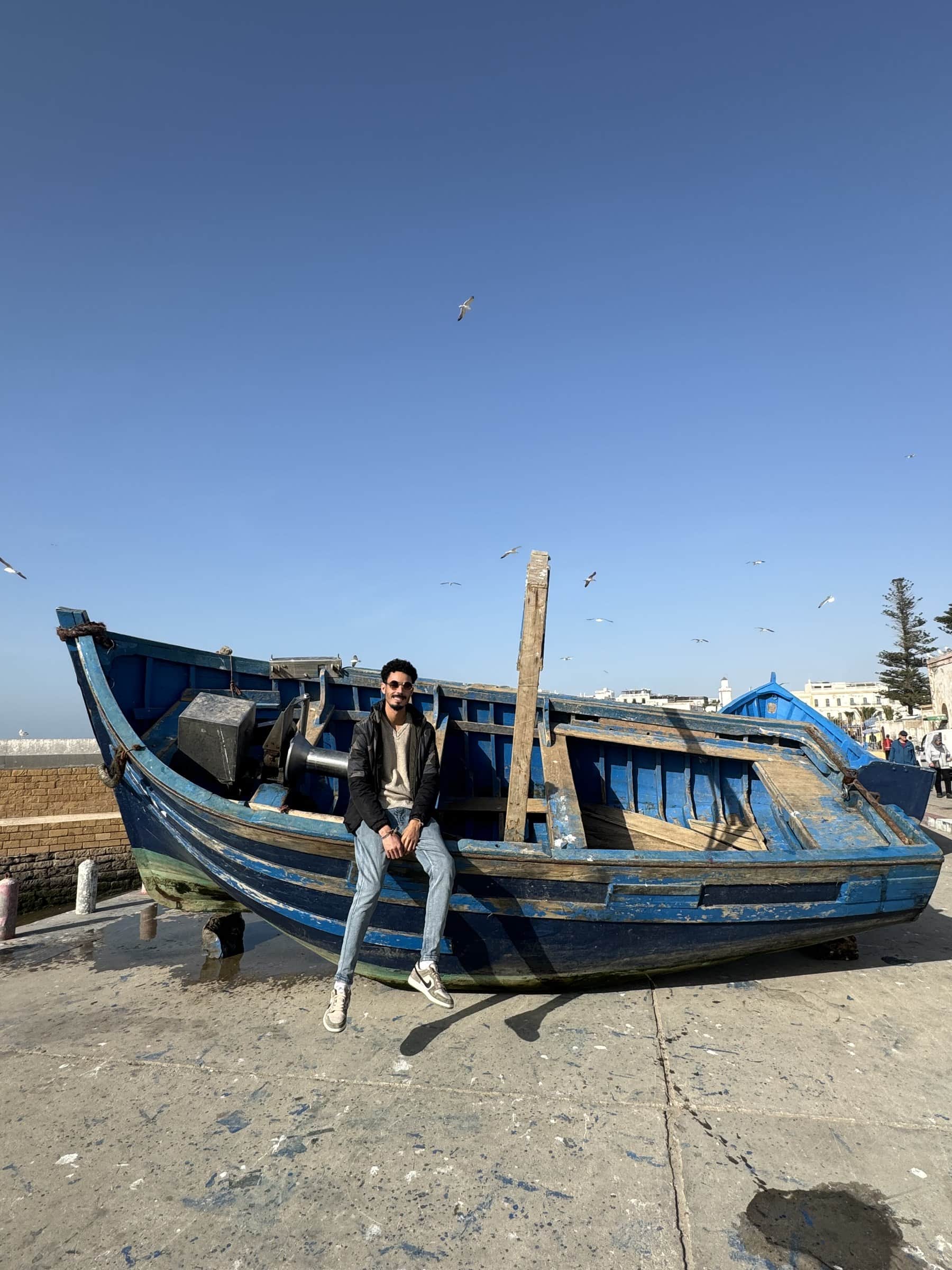 Hombre apoyado en barca de pesca azul tradicional en el puerto de Essaouira con gaviotas volando