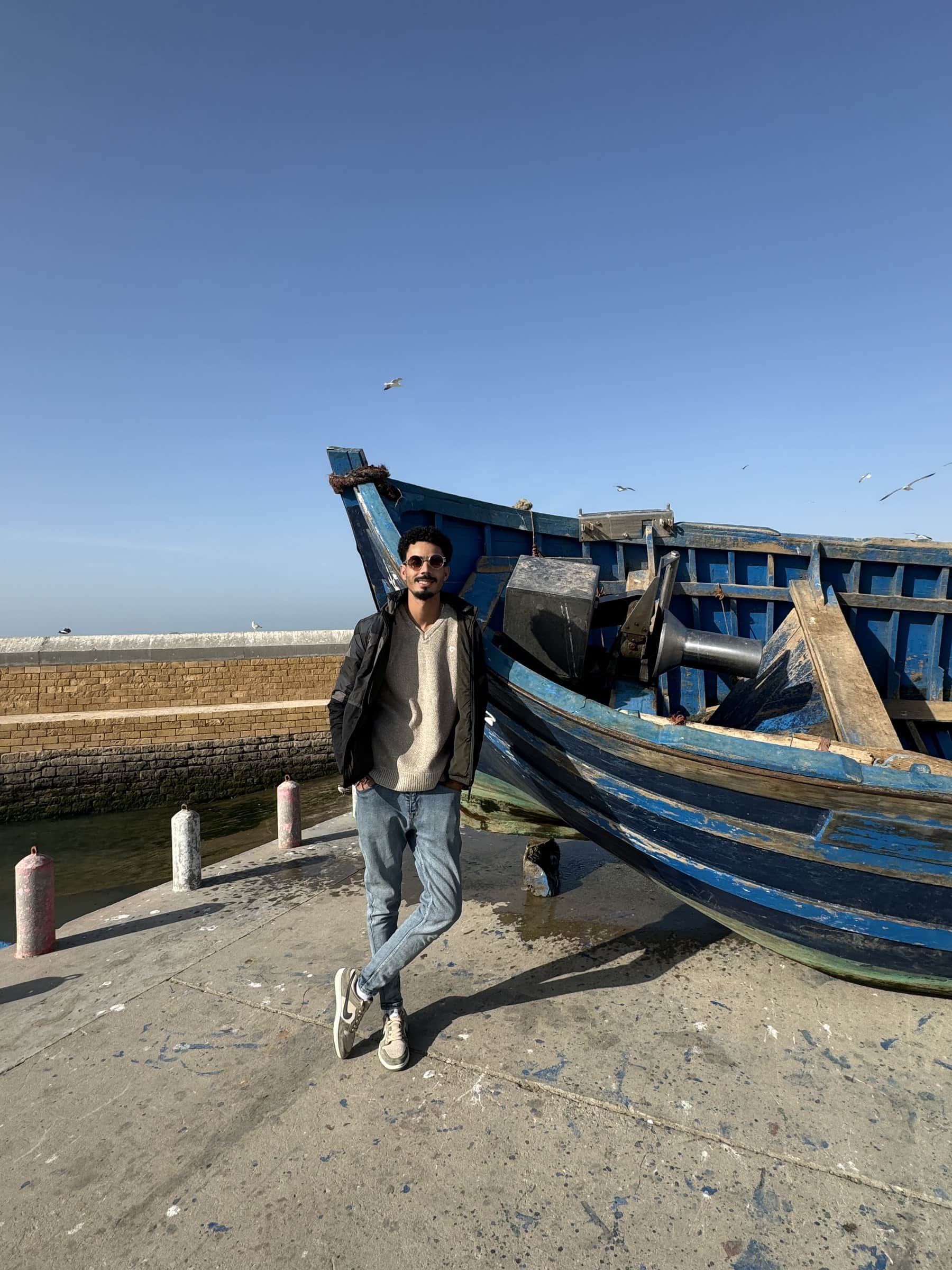 Joven posando junto a barcos de pesca azules en el puerto histórico de Essaouira, Marruecos