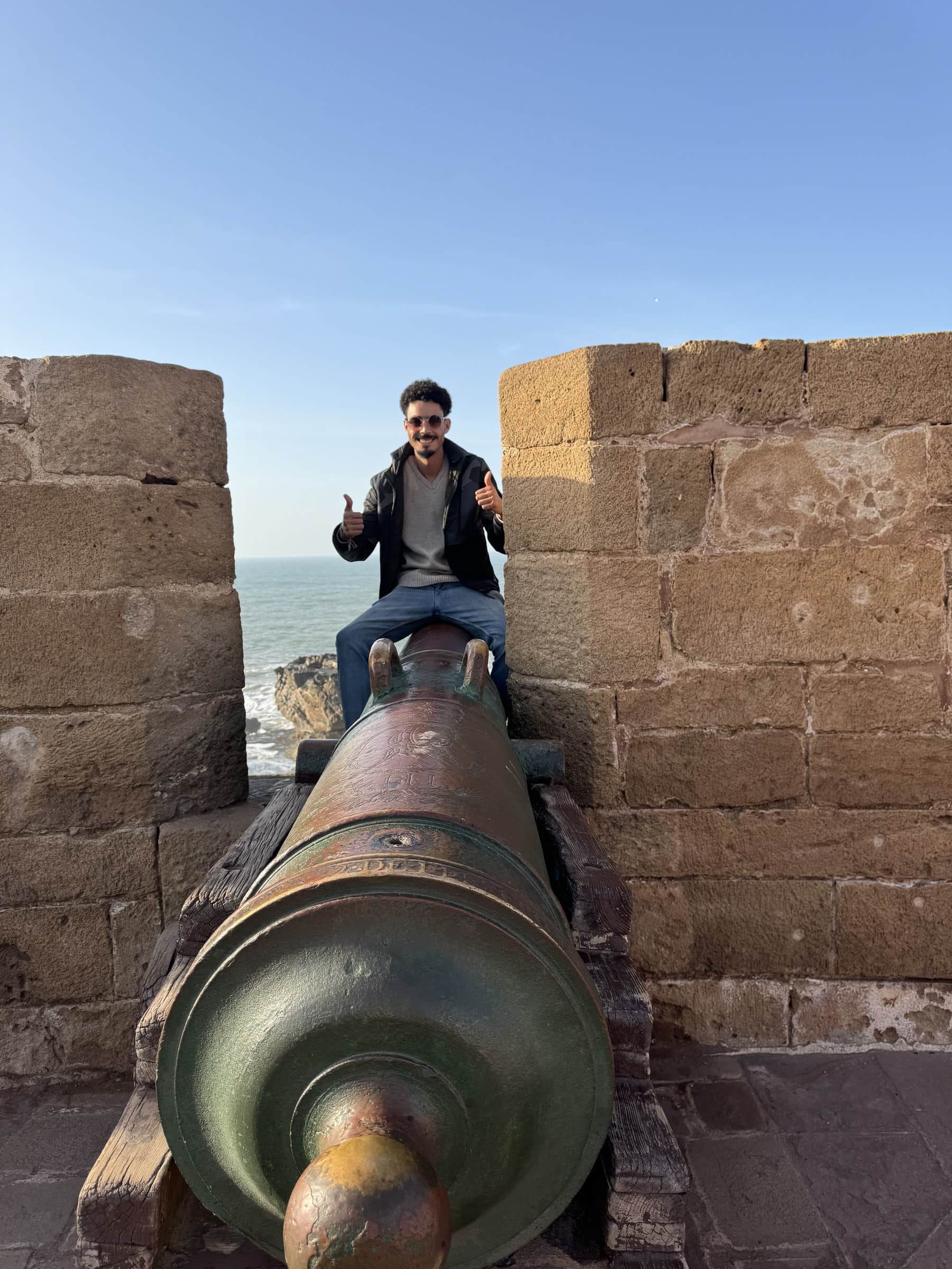 Joven sentado sobre un cañón histórico en las murallas de Essaouira con el océano Atlántico al fondo