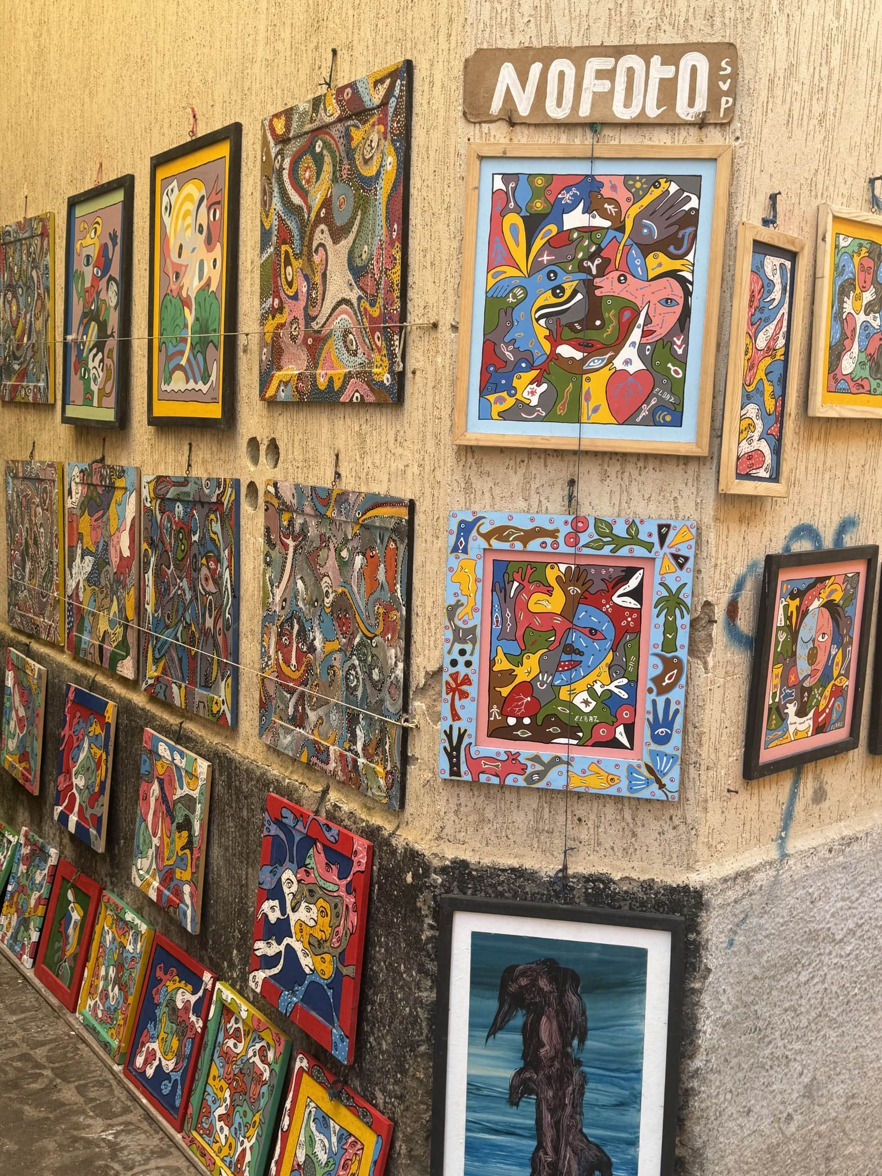 Pinturas de arte popular marroquí expuestas en una pared de la medina con cartel No Foto