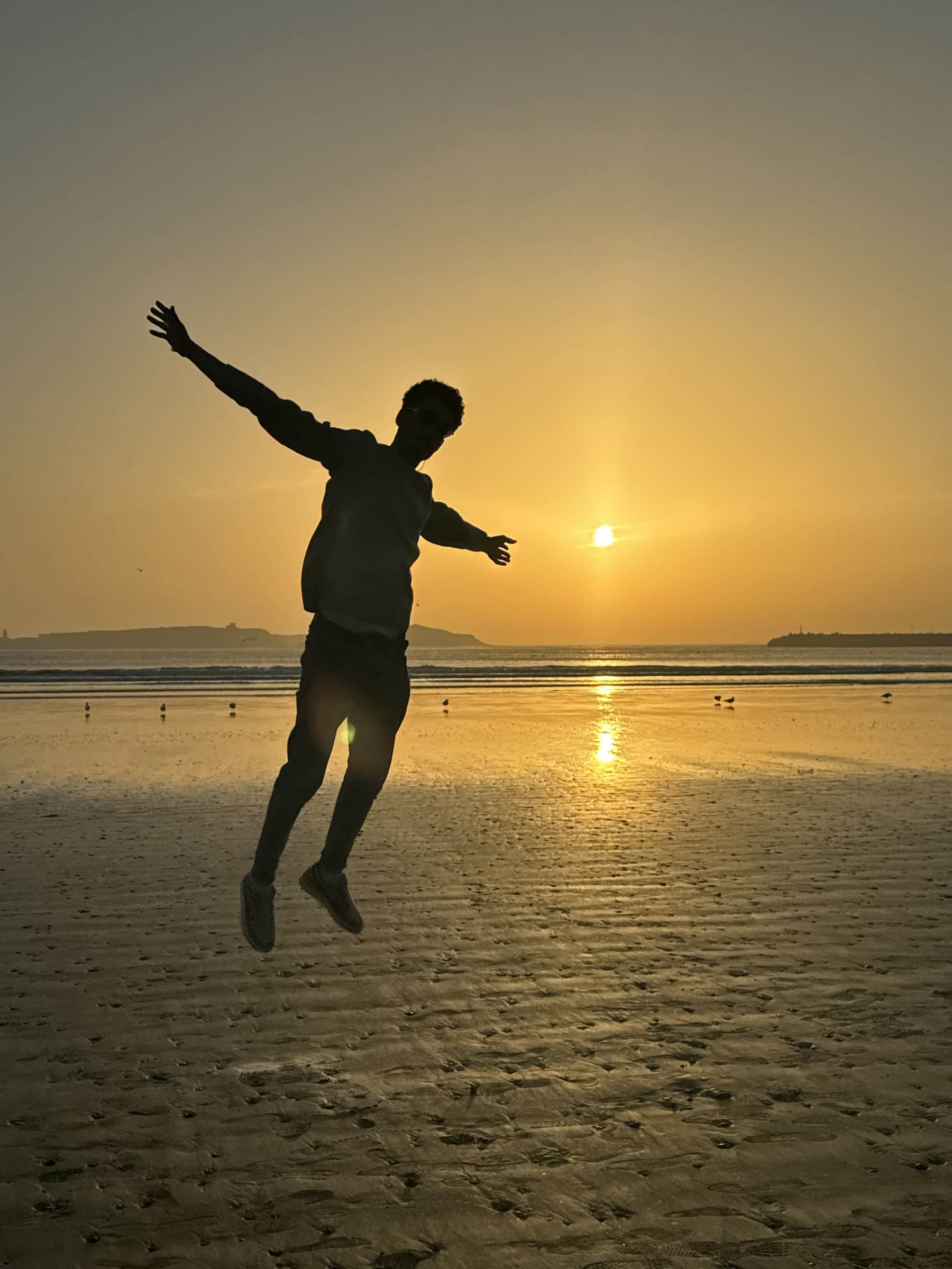 Silueta de persona saltando en la playa al atardecer en Essaouira, Marruecos, con el sol reflejado en el agua
