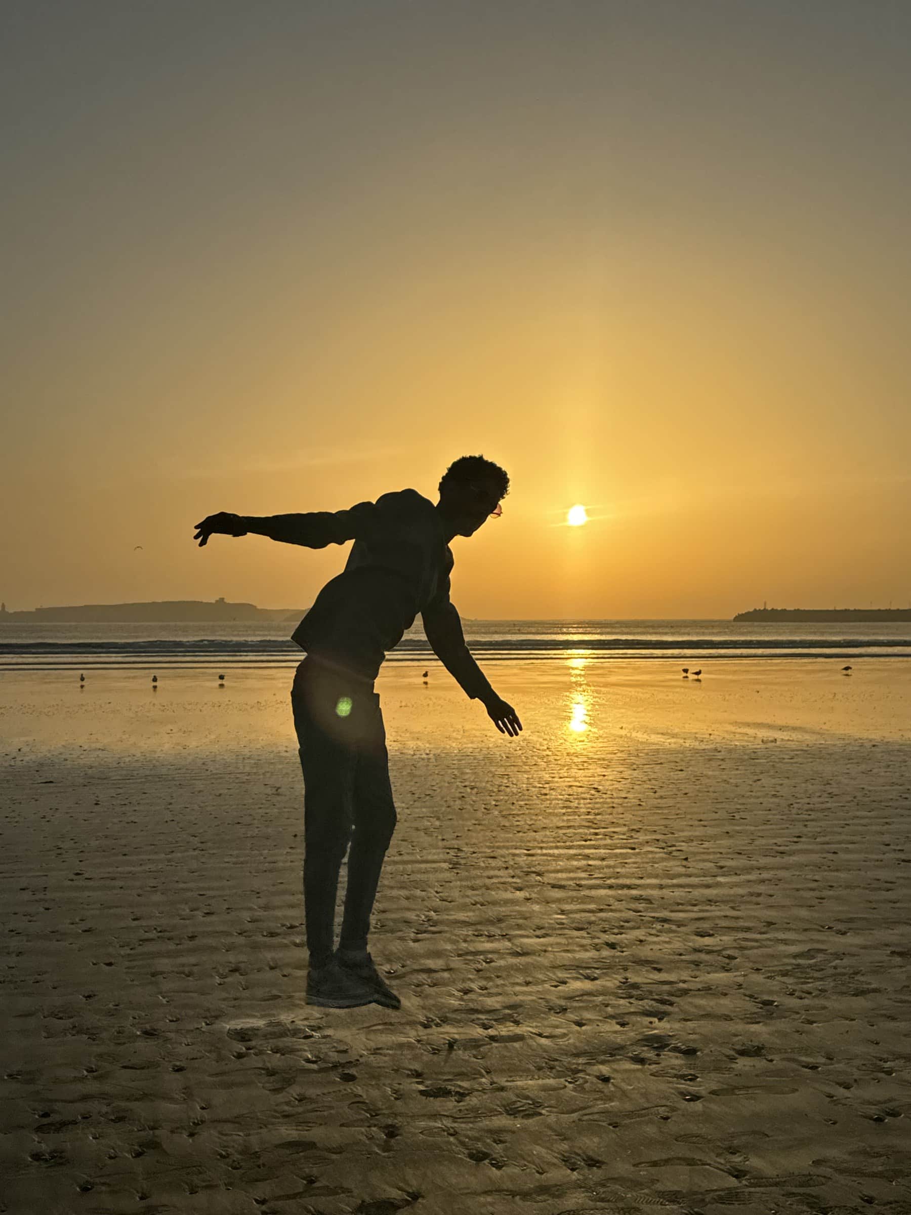 Silueta de persona en playa atlántica de Marruecos al atardecer con cielo dorado y sol en el horizonte