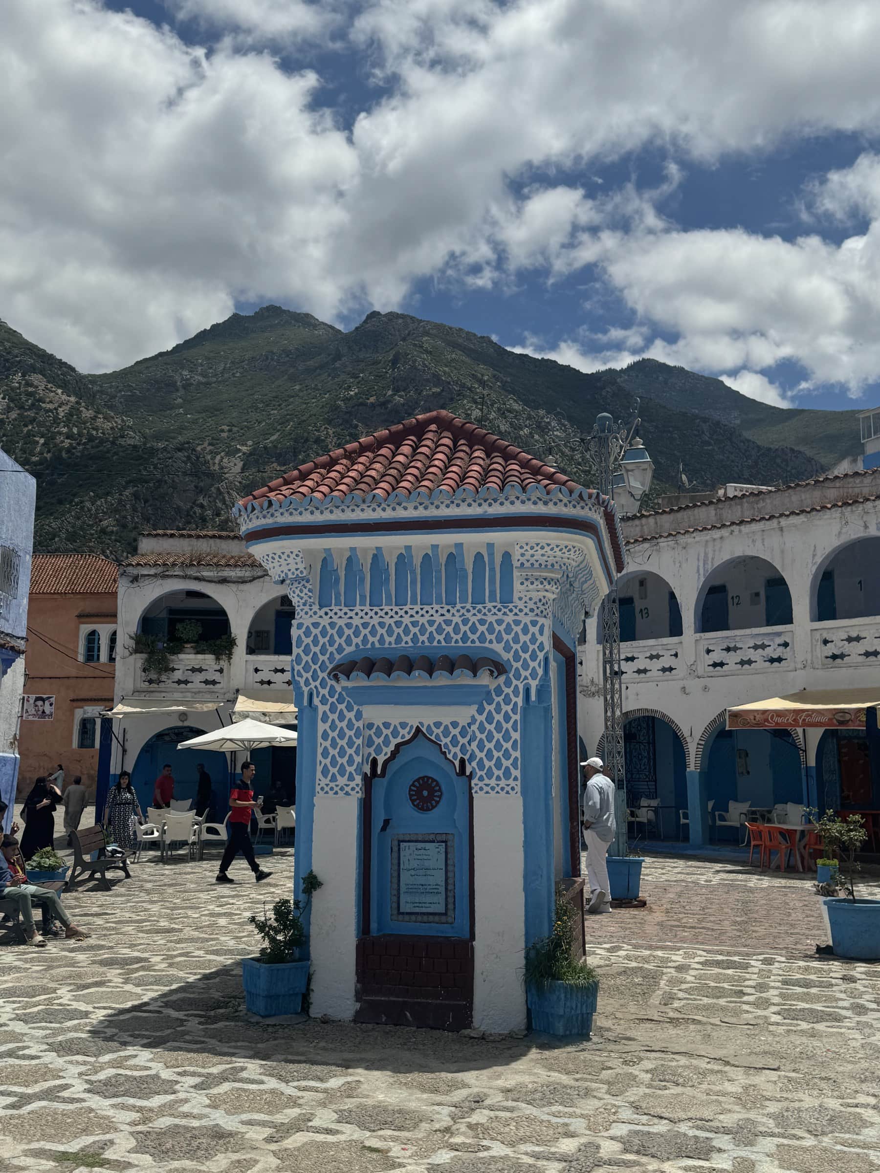 Plaza Uta el-Hammam de Chefchaouen con fuente azul y blanca, montañas del Rif al fondo, Marruecos