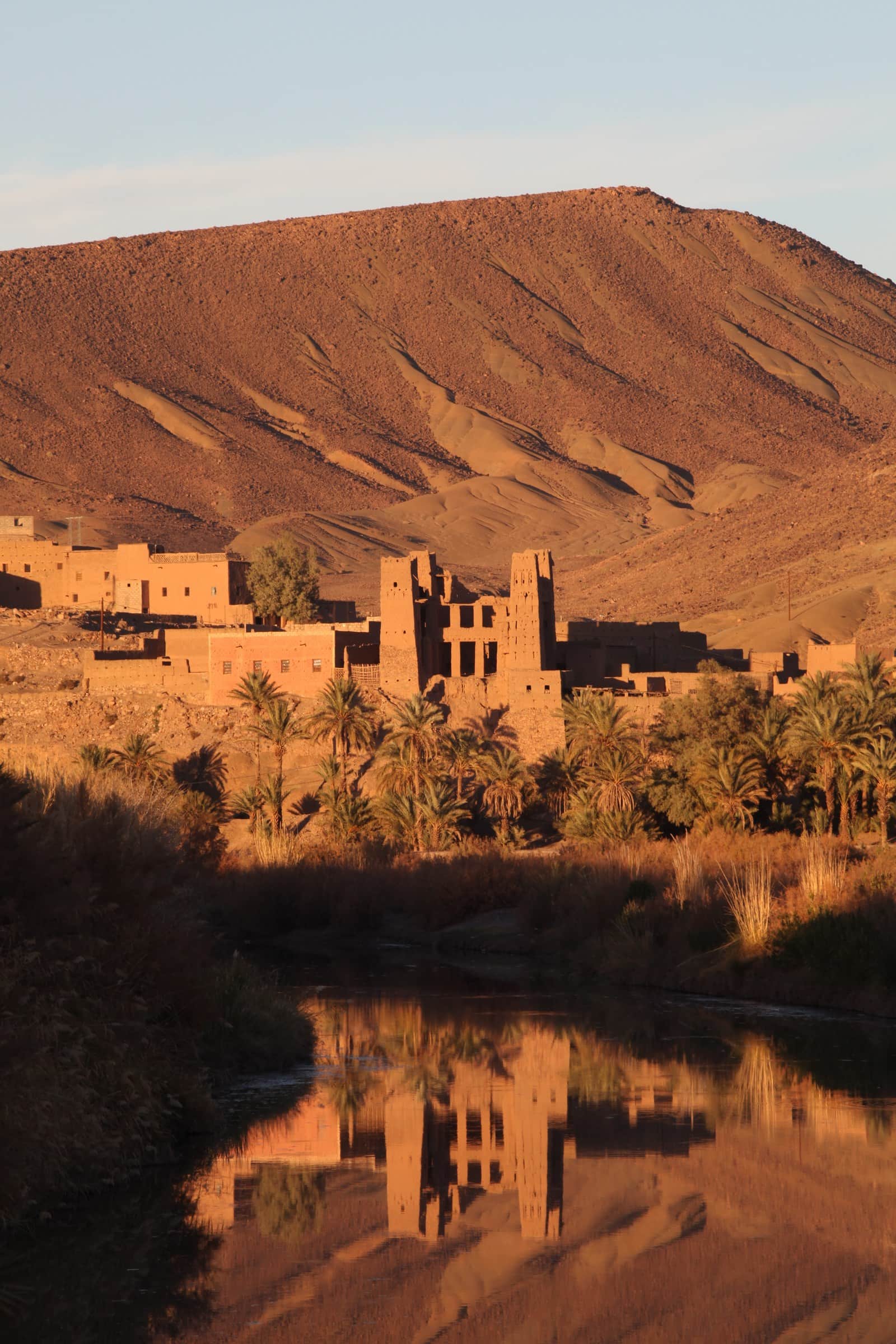 Kasbah de barro reflejada en el río con palmeras y montañas ocres al atardecer en el Valle del Draa, Marruecos