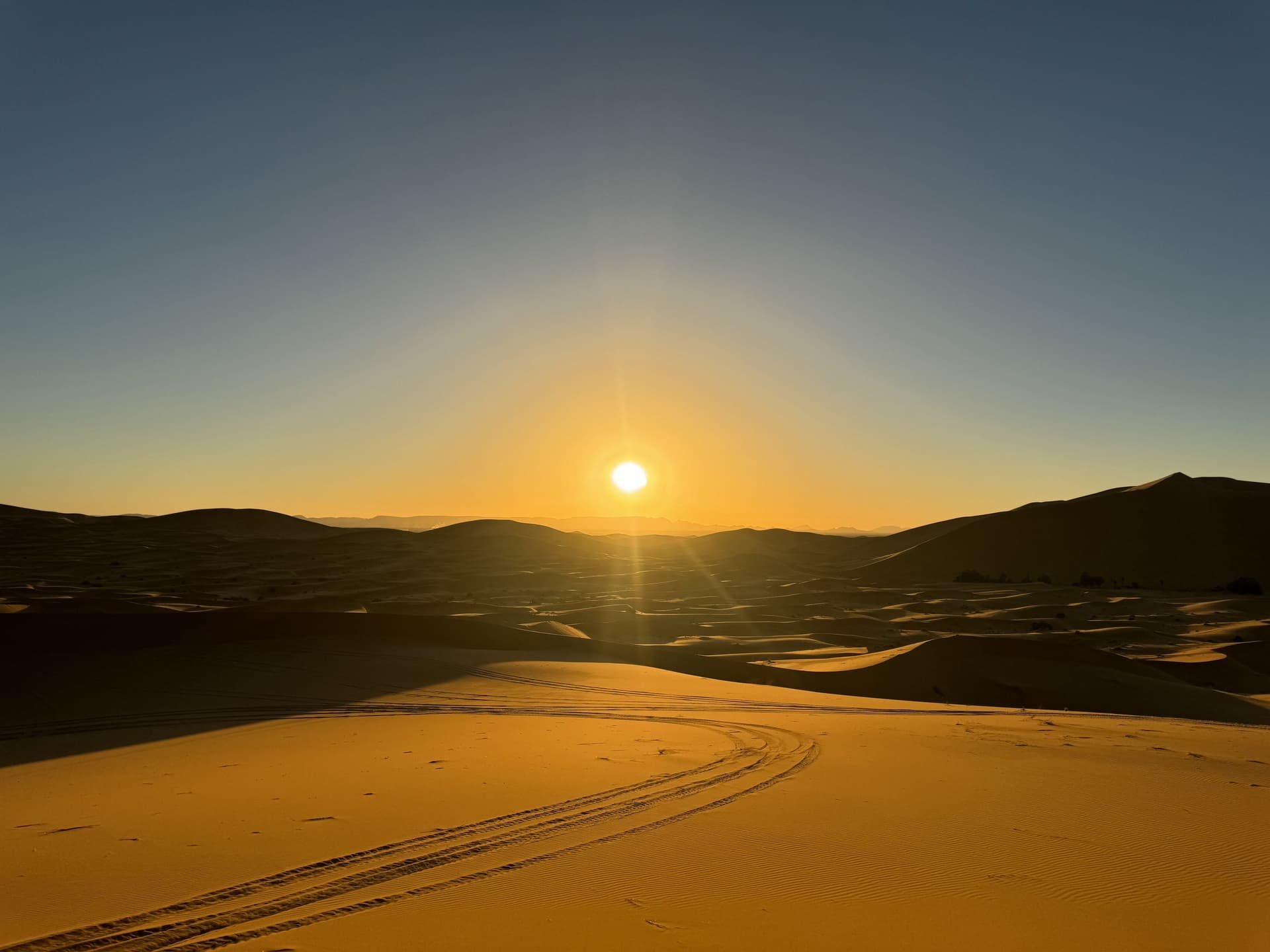 Amanecer dorado sobre las dunas del Erg Chebbi en Merzouga, con huellas de 4x4 en la arena del Sahara