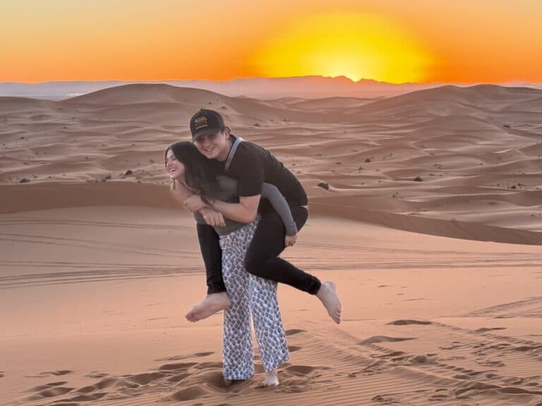 Pareja jugando y riendo sobre las dunas del desierto de Merzouga al atardecer con cielo naranja