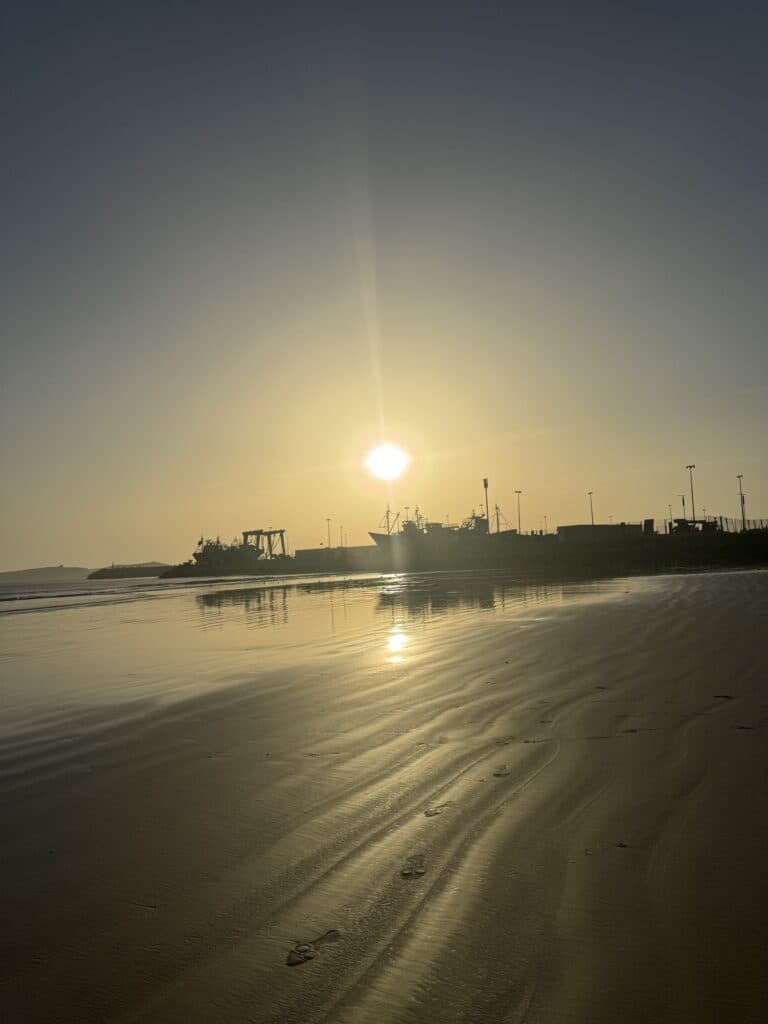 Atardecer dorado en la playa de Essaouira con silueta del puerto pesquero reflejada en la arena mojada