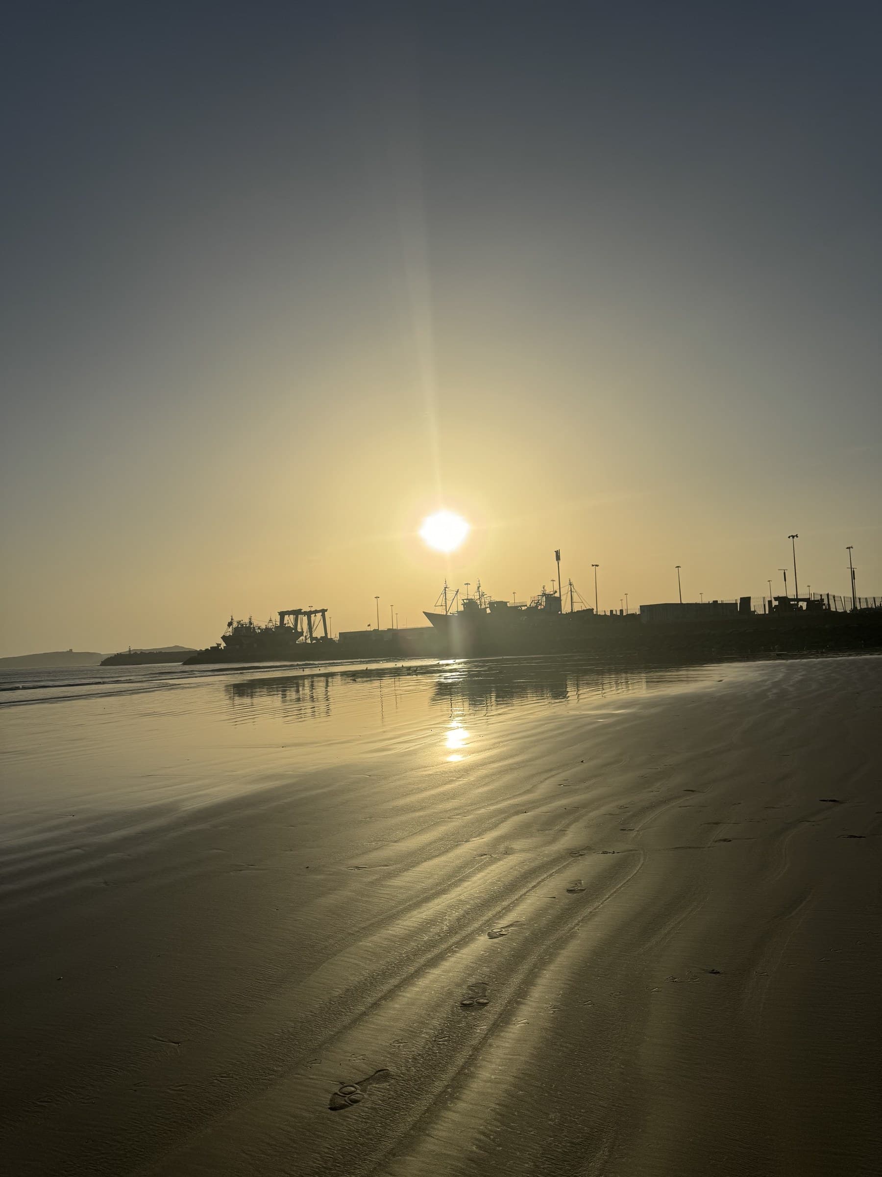 Atardecer dorado en la playa de Essaouira con silueta del puerto pesquero reflejada en la arena mojada