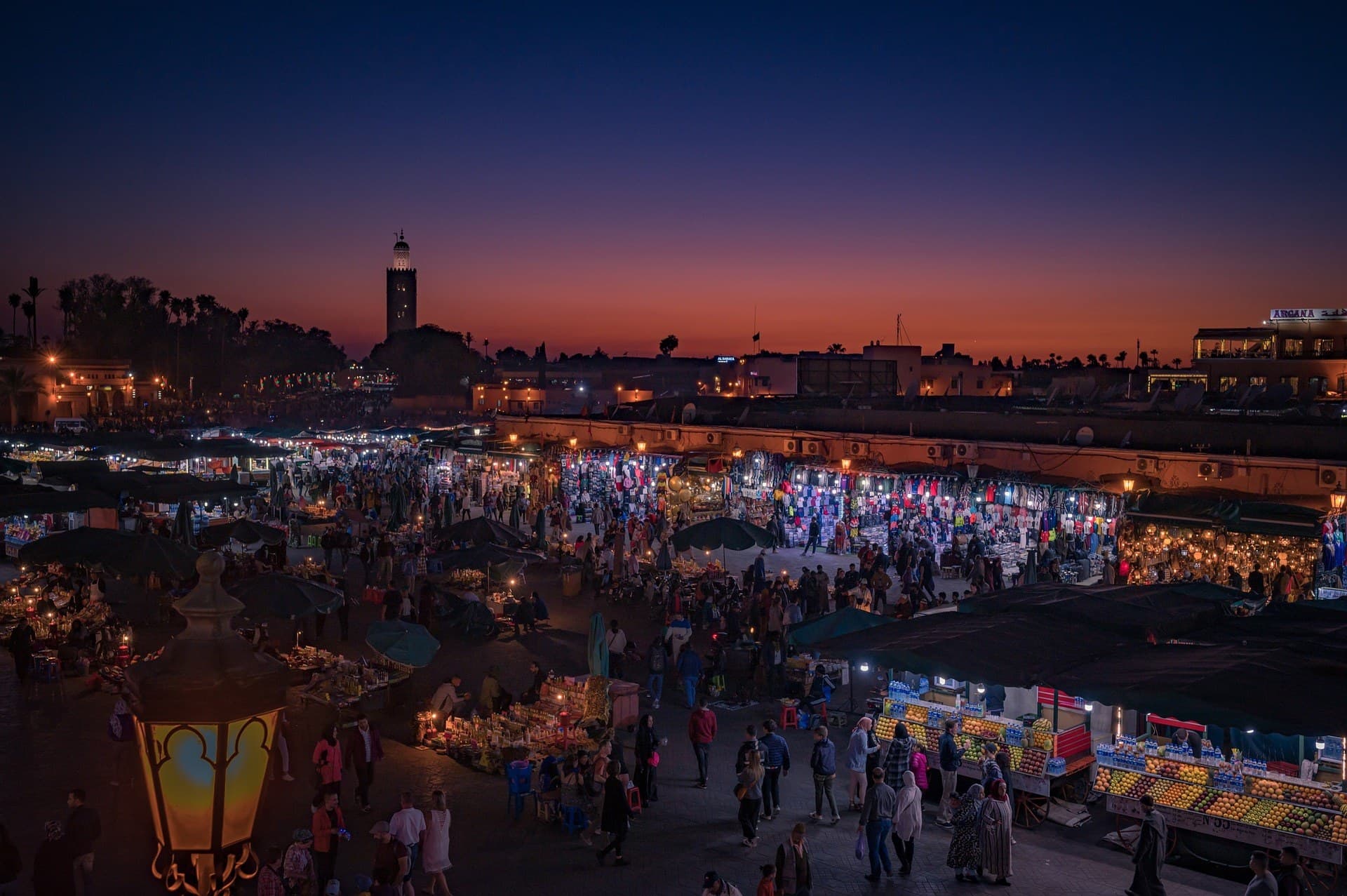 Plaza Jemaa el-Fna de Marrakech al atardecer con puestos iluminados, multitud y minarete Koutoubia al fondo