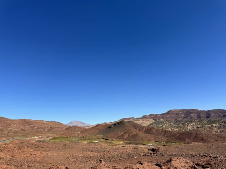 Valle árido del Alto Atlas marroquí con tierras rojizas, vegetación en el fondo y cielo azul intenso