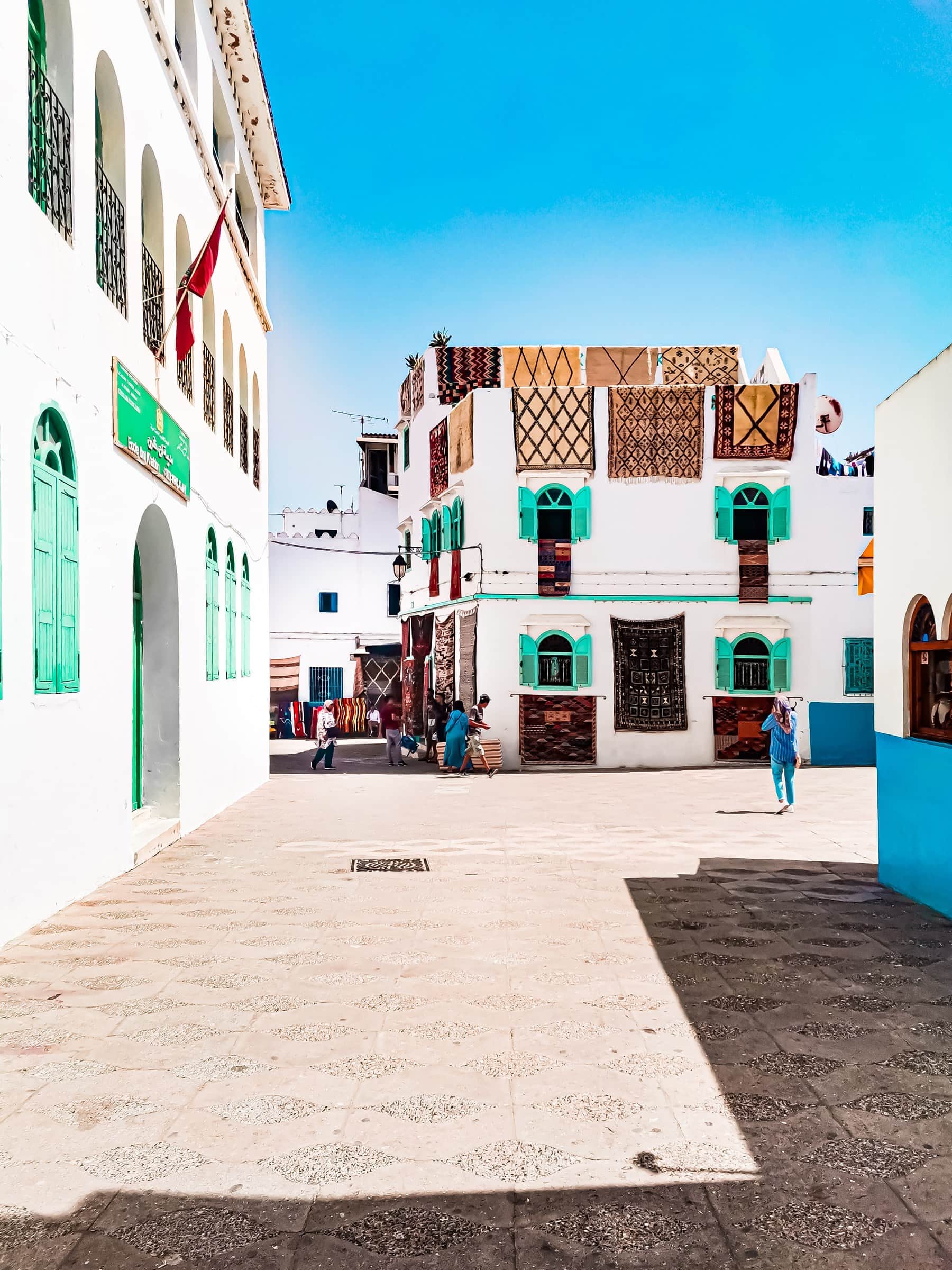 Plaza de la medina de Asilah con edificios blancos, contraventanas verdes y alfombras bereberes colgadas en las fachadas